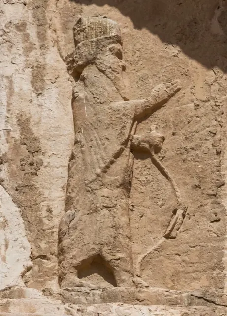A worn stone relief of a bearded Persian king in a tall crown, raising one hand and holding a bow